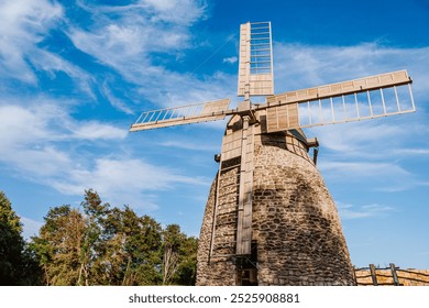 Old stone windmill stands against a clear blue sky with gentle clouds. The windmill wooden blades and stone structure showcase traditional craftsmanship  - Powered by Shutterstock