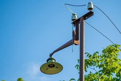 An Old Street Lamp On A Rusty Pole With A Broken Bulb And Torn Wires Against A Background Of Green Leaves And Blue Sky