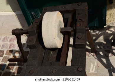 Old Style Round Grindstone Wheel. Knife Sharpening Tool Set In Wood Frame. Vintage Equipment. Rusty Iron Axis. Stucco Wall Background. Decorative Piece At Street Storefront. Rusty Steel Metal Spindle