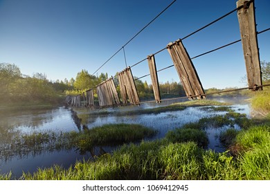 Old Suspension Bridge Over The River In The Forest. The Rope Construction Is Broken And Skewed, It's Dangerous To Cross. Spring Landscape With Bright Juicy Paints Against The Blue Sky.