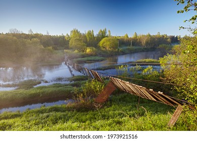 Old Suspension Bridge Over The River In The Forest. The Rope Construction Is Broken And Skewed, It's Dangerous To Cross. Spring Landscape With Bright Juicy Paints Against The Blue Sky.