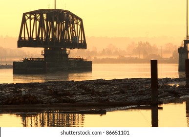 An old, swing span, rail bridge over the Fraser River is open to allow ship traffic. A log boom is tied to pilings in the foreground. Early morning, just before sunrise. British Columbia, Canada.