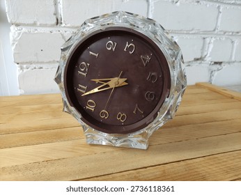 Old table clock with glass case on wooden table against a white brick wall