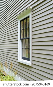 Old Tan Colored House With White Trim And Black Wood Window Frame Spacers. The Glass In The Window Is Wavy And Reflects The Sun. The Exterior Wall Is Made Of Vintage Beige Cape Cod Clapboard Siding.