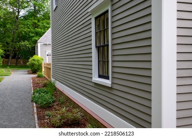 Old Tan Colored House With White Trim And Black Wood Window Frame Spacers. The Glass In The Window Is Wavy And Reflects The Sun. The Exterior Wall Is Made Of Vintage Beige Cape Cod Clapboard Siding.