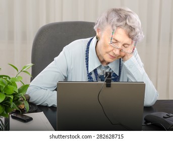 Old Teacher Is Falling Asleep In Front Of Laptop Screen During Lesson On-line. Grey Haired Old Woman Is Wearing Blue Blouse And Dark Blue Stone Necklace. The Glasses Will Fall Off Her Face Soon.