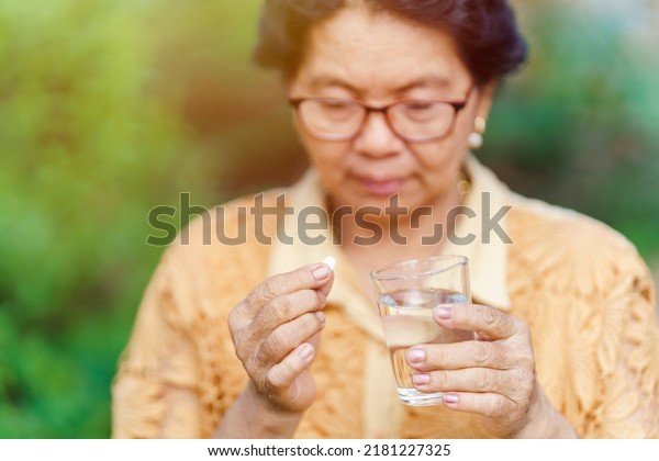 An old Thai woman sits and looks at the pill in\
her hand with a glass of water to eat and relieve sickness. Health\
care concept.