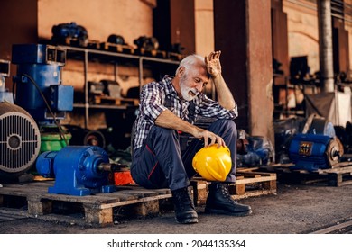 An old, tired bearded factory worker in overalls is sitting on the pallet and taking a break from hard work. He is wiping sweat from his forehead. - Powered by Shutterstock