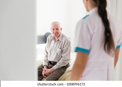 The Old Tired Man Sitting On A Bed In The Room At Home And Young Nurse Entering In A Room