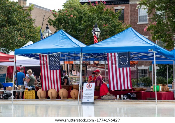 Old Town Alexandria, Virginia / USA - July
11 2020: Vendors set up stalls with colonial American flags at the
farmers market. Cautious customers shop with blue medical masks due
to the coronavirus.