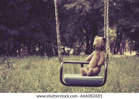 Old toy teddy bear sitting on swing in park, retro style, toned effect
