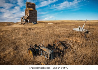 An old tractor is laying in a field next to a grain silo. The tractor is rusted and the silo is in a state of disrepair. Concept of abandonment and neglect, as the old farm equipment - Powered by Shutterstock