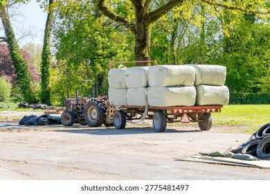 Old tractor pulling a flatbed trailer loaded with white wrapped hay bales across a sunny rural farmyard with trees and tires