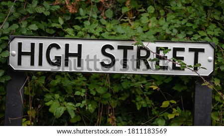 Old traditional English high street sign in small rural village surrounded by dark green foliage. Shops highstreet shopping town centre.
