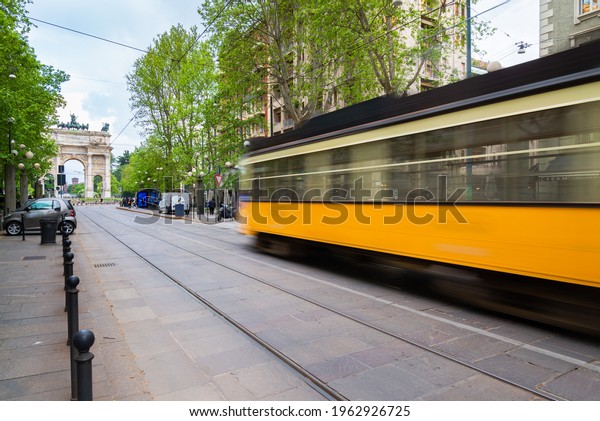 Old traditional tram in\
motion blur arriving at the station with passengers waiting, Milan,\
Italy. Arco della Pace (meaning: Arch of the Peace) in the\
background.