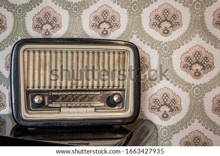 An old transistor radio, with knobs and buttons for manual tuning. In the background a vintage wallpaper. Ancient object, worn and ruined by time.