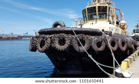 Old tugboat moored at berth in maritime port with industrial seascape on the background. Used black tires at shipboard of tug ship in port. Maritime transportation scene.