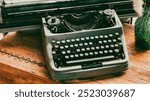 An old typewriter sits on a wooden table. The typewriter is black and white and has a vintage look. The table is made of wood and has a vase on it. The vase is green and has a floral design
