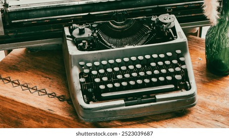 An old typewriter sits on a wooden table. The typewriter is black and white and has a vintage look. The table is made of wood and has a vase on it. The vase is green and has a floral design