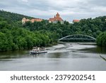 The old Veveri castle on the hill above the Brno dam, over which a bridge leads and a cruise ship with tourists floats on the water.