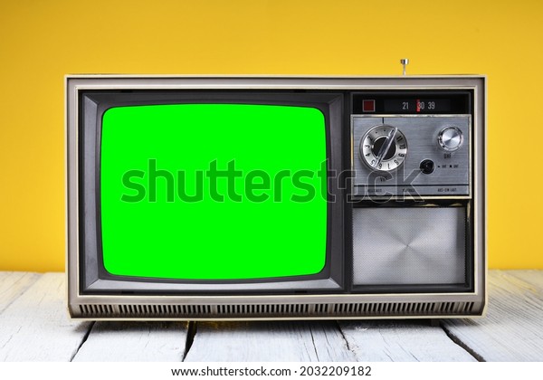 An old vintage 1970s TV with a green screen for\
adding video stands on a wooden table against a yellow background.\
Vintage TVs 1980s 1990s 2000s.\

