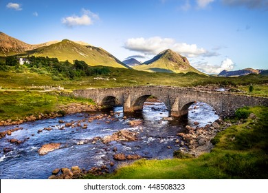 Old vintage brick bridge crossing river in Sligachan, Isle of Skye, Scotland with scottish landscape, vegetation, hills and mountains with fresh, blue water and sunny sky