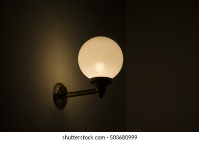 An old, vintage light bulb in a round lampshade, fixed to the wall with a golden light fixture, with paint on it. The image is underexposed to emphasize the dark, mysterious mood of the setting. - Powered by Shutterstock