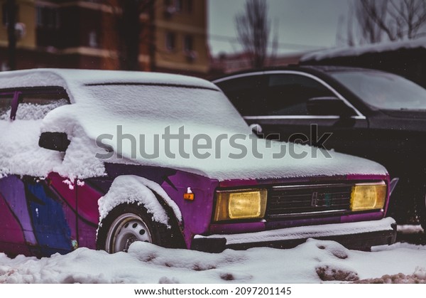 Old\
vintage Russian car covered in snow with headlights on in a moody\
dark atmospheric setting. Contrasted with new\
cars.