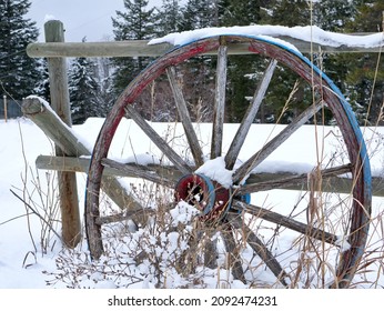An old wagon wheel propped up against a wooden fence, covered in winter snows.