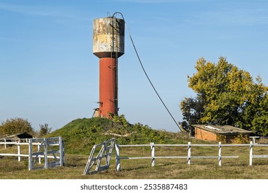 Old water tower in the countryside. A rusty, outdated water tower stands in an open field with a damaged fence and a small brick shed next to it. The scene evokes rural neglect and rural charm - Powered by Shutterstock