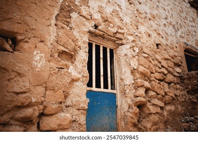 An old, weathered stone wall with a rustic blue door and barred window. The texture of the stones is rough, and the paint on the door is peeling, suggesting age and neglect. - Powered by Shutterstock