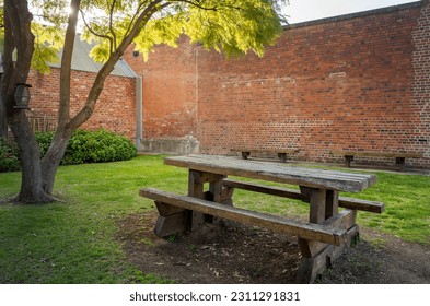An old and weathered wooden picnic bench and table under a tree in a courtyard with classic red brick walls in warm natural sunlight. - Shutterstock ID 2311291831