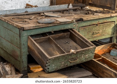 Old, weathered workbench with open drawer and scattered tools including metal plates and a saw blade, set in a rustic workshop or garage, captured in natural light with gritty detail.