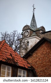 Old White Church Tower Near Old City Houses. Photo Taken In Europe, Latvia, On A Cloudy, Warm Winter Day.