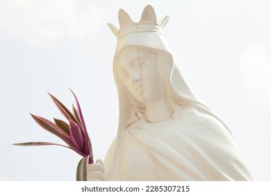 Old white sculpture of a saint woman wearing a crown and holding a bouquet. Catholic holy vintage statue against the light sky
