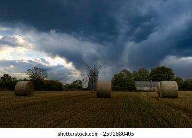 


An old windmill under a dramatic stormy sky with a field and hay bales