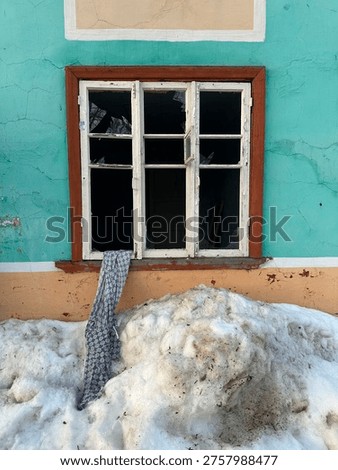 Old window in a turquoise wall with deep snow and a wooden shovel outside.