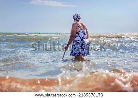 An old woman in a bathing suit with a cane went into the sea knee-deep, rear view