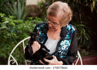 An Old Woman, With A Black Cat, Sitting In A Chair In A Garden.