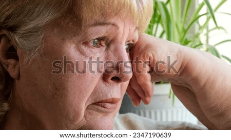 Old woman crying at the window. Face of depressed elderly woman at home close up. Upset senior elder grandma grieving, weeping, suffering from anxiety, grief, sorrow or disease