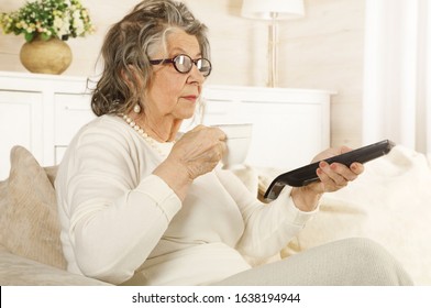 An Old Woman Is Drinking Tea With A TV Remote In Her Hand Sitting On The Sofa Indoors
