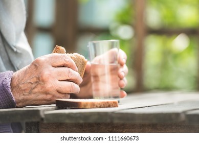 The Old Woman Drinks Water And Eats Bread At An Old Wooden Table In A Garden