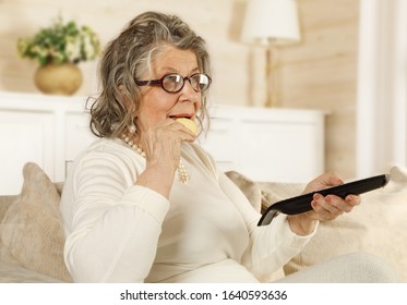 An Old Woman Eating Cookies With A TV Remote In Her Hand Sitting On The Sofa Indoors
