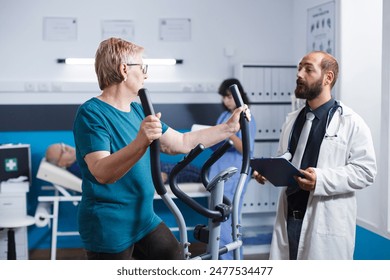 Old woman exercises on electronic bicycle while recovering from an injury. Male physician with a tablet provides physiotherapy support to elderly patient who does a workout on stationary bike. - Powered by Shutterstock