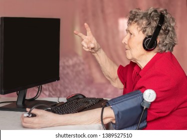 An old woman with gray hair and headphones on her head looks at the computer screen. Grandmothers at the computer are happy with their victory in a computer game.
