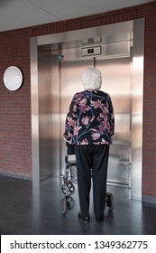 Old Woman With Grey Hair Walking With Her Walker, In Nursing Home With Elevator. 