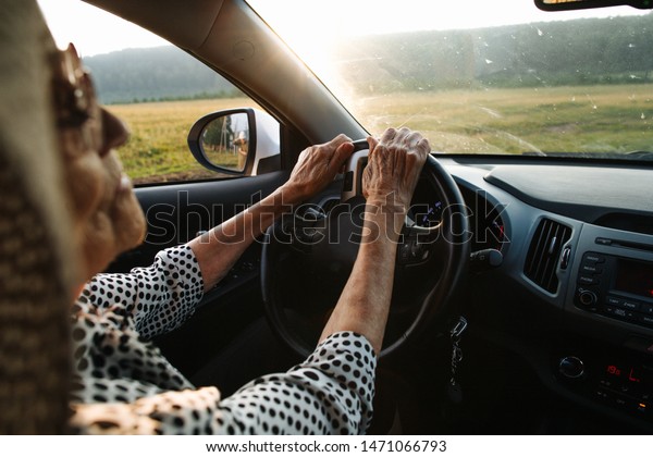 Old woman in a\
hat driving a car rides on the field. Holds hands on wheel. Looks\
carefully at the road.\
Sunset