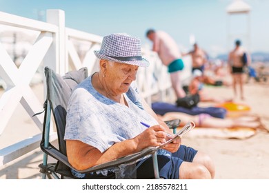 An old woman in a hat sits in a tourist chair on the beach and solves crossword puzzles on a sunny day - Shutterstock ID 2185578211