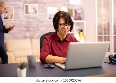 Old Woman In Her 60s Using A Modern Laptop In Her Cozy House Late In The Evening