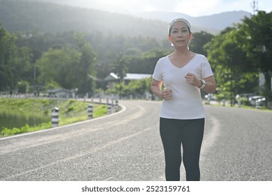 An old woman jogs on a road surrounded by nature, with mountains in the background, showcasing her dedication to staying fit and active at any age, Age is No Barrier, Empowerment Through Fitness. - Powered by Shutterstock